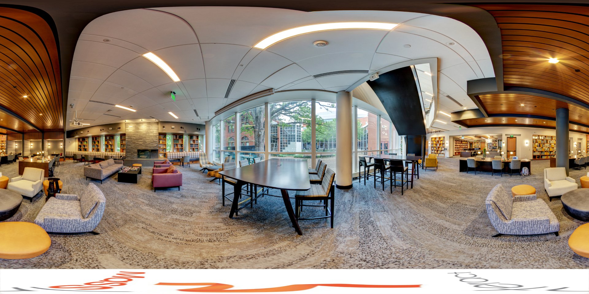 A panoramic view of a cozy, modern library space with comfortable seating, including sofas and chairs, surrounding a fireplace. Large windows provide natural light and a view of the trees outside. The room includes long tables for studying and bookshelves lining the walls.